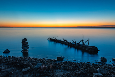 Rocks stacked by a dead tree near a lake shore at sunset